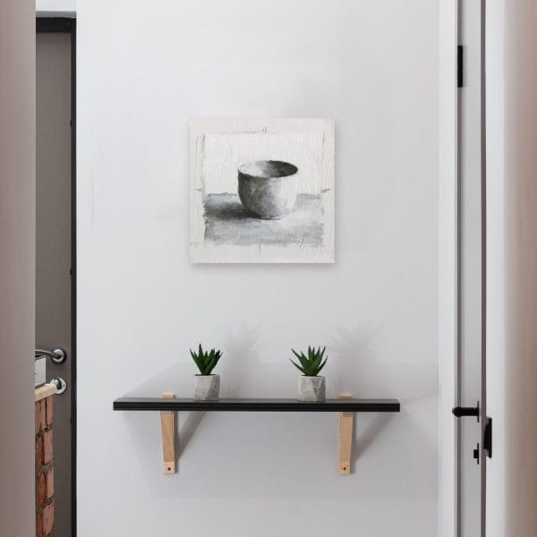 Monochromatic oil painting of a white cup on a white background with black and grey shadows displayed on a wall.