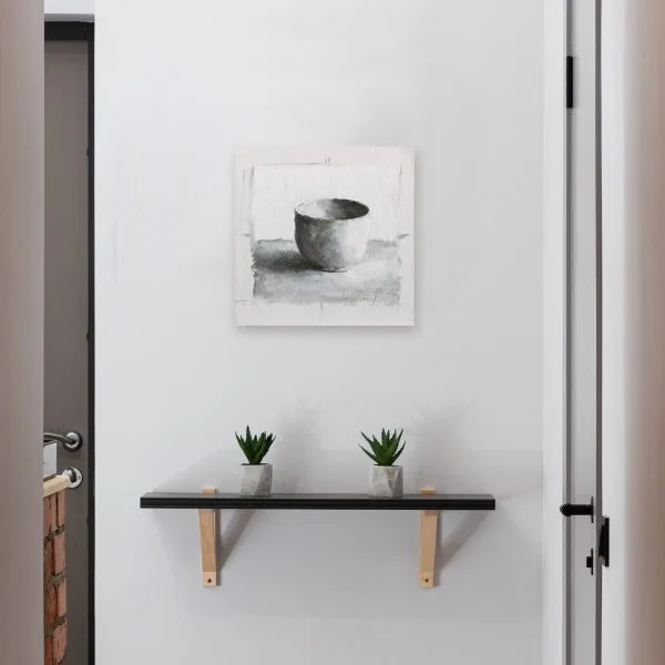 Monochromatic oil painting of a white cup on a white background with black and grey shadows displayed on a wall.