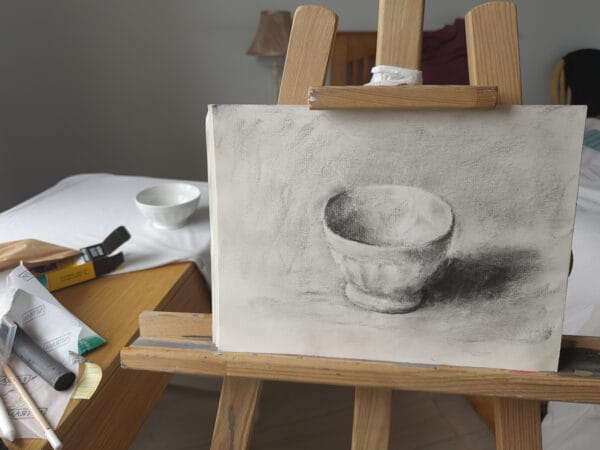 Charcoal drawing on paper of a white bowl display in an easel with the still life setup in the background.